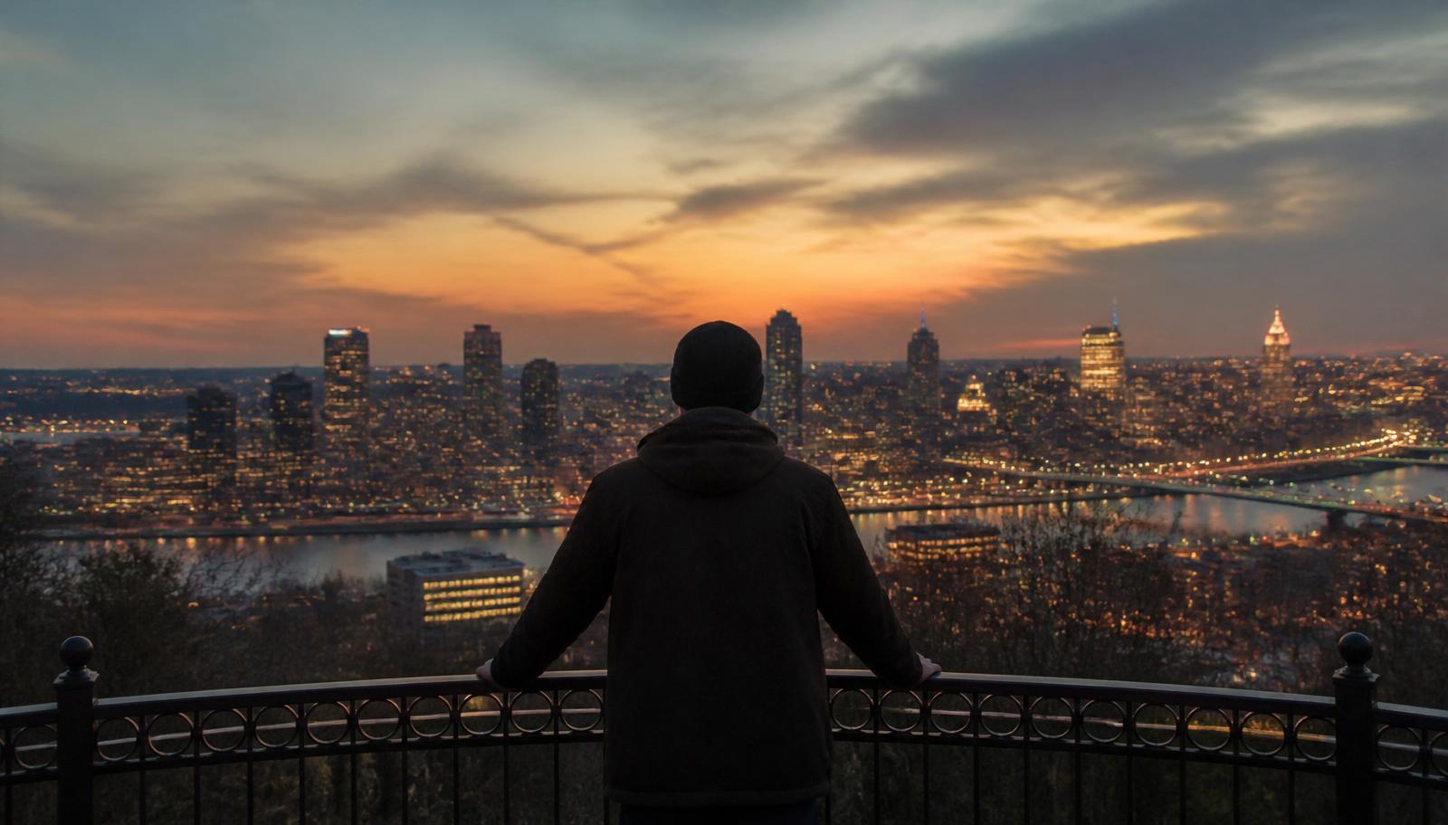 A lone figure standing at a park overlook at dusk, quietly watching over a city skyline as the lights come on — the vigilance of cybersecurity as a human mission